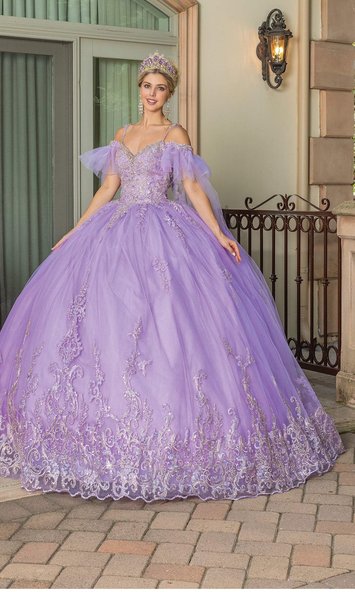 A woman stands outdoors on a stone patio in front of a building, smiling with arms slightly raised, wearing Quinceanera Dress 1713 By Dancing Queen—a sparkling lavender off-the-shoulder gown with ruffle sleeves and a tiara.